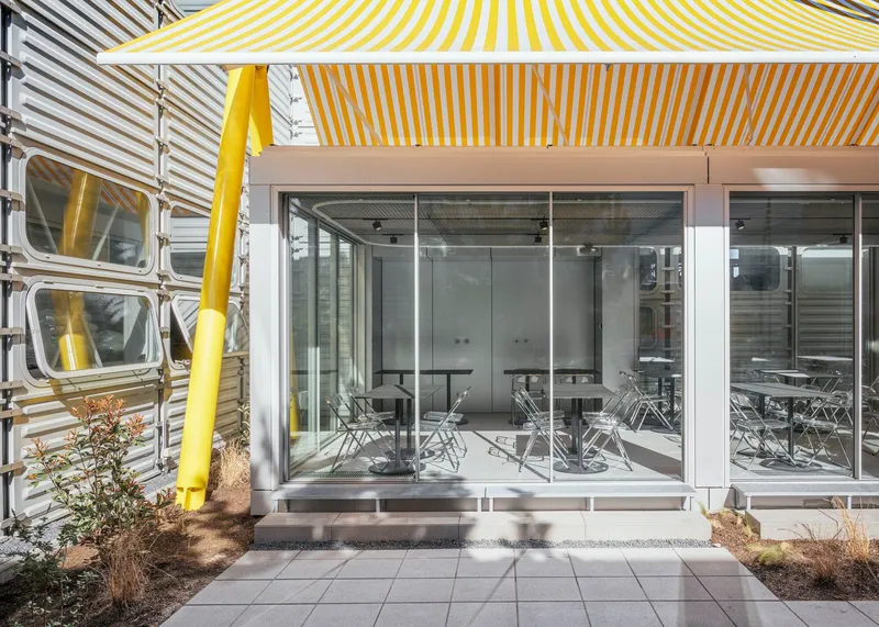Glass entrance with sliding doors under yellow and white awning framed by yellow structural columns and planting