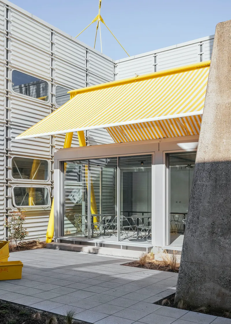 Covered entry threshold with yellow striped fabric canopy, concrete column, and corrugated metal facade under blue sky