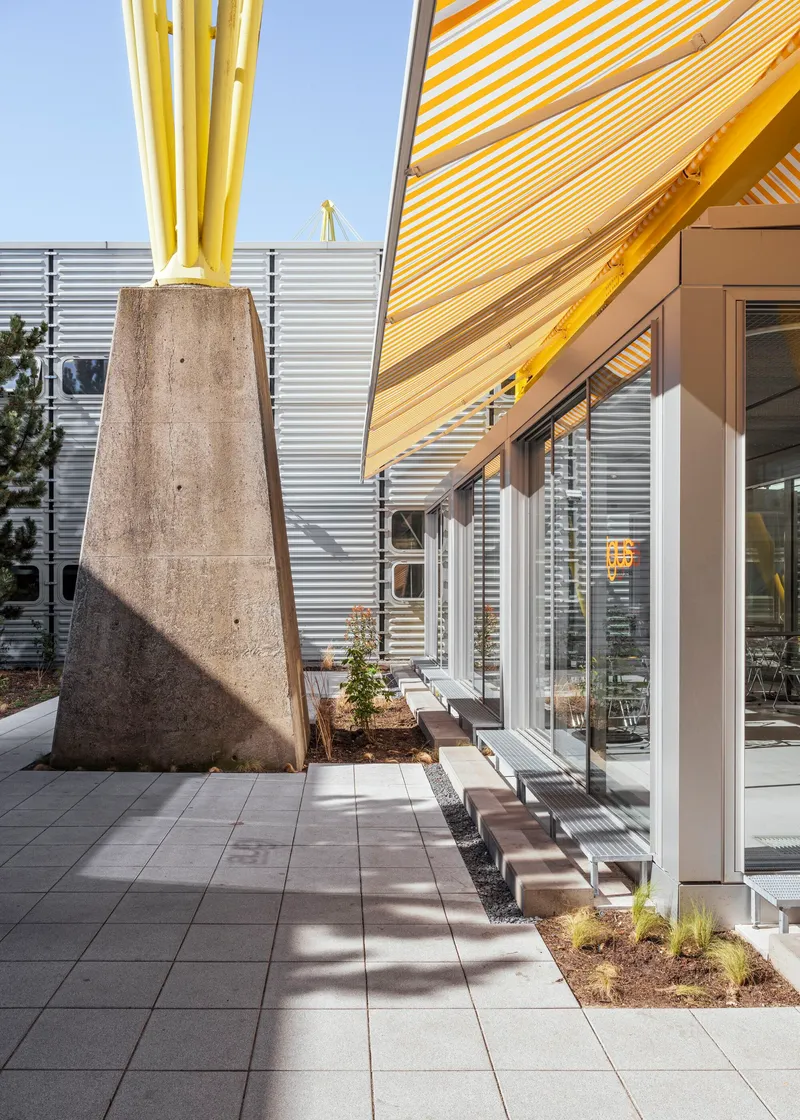 Tapered concrete column supporting yellow striped canopy beside corrugated metal facade and paved courtyard
