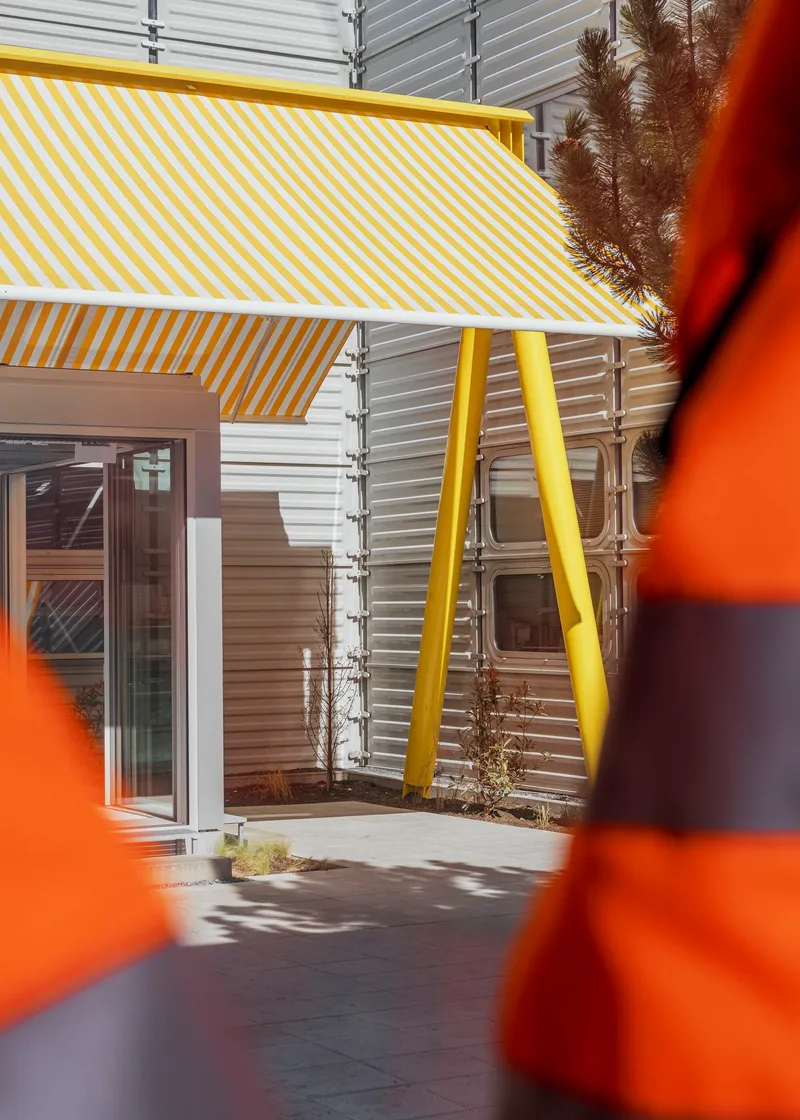 Courtyard view through orange bollards showing yellow canopy and planted beds along corrugated facade