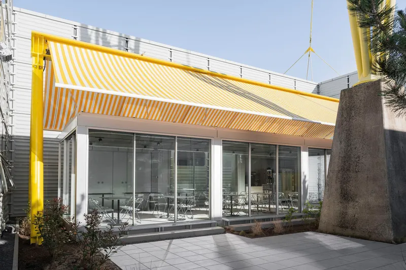 Glass-walled dining pavilion with yellow striped canopy flanked by concrete column and evergreen trees
