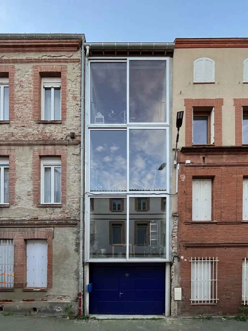 Narrow glass facade with white frames and a blue garage door inserted between weathered brick rowhouses