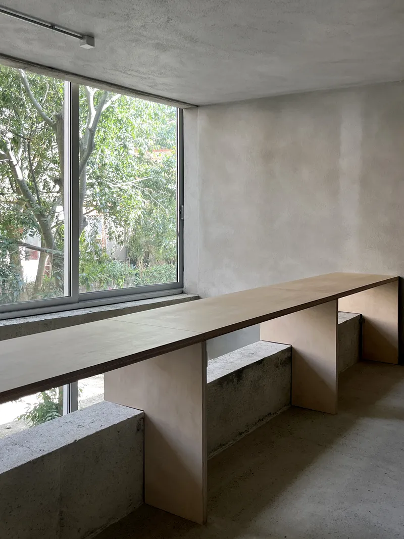 Built-in plywood desk and concrete seating bench beside a window overlooking trees