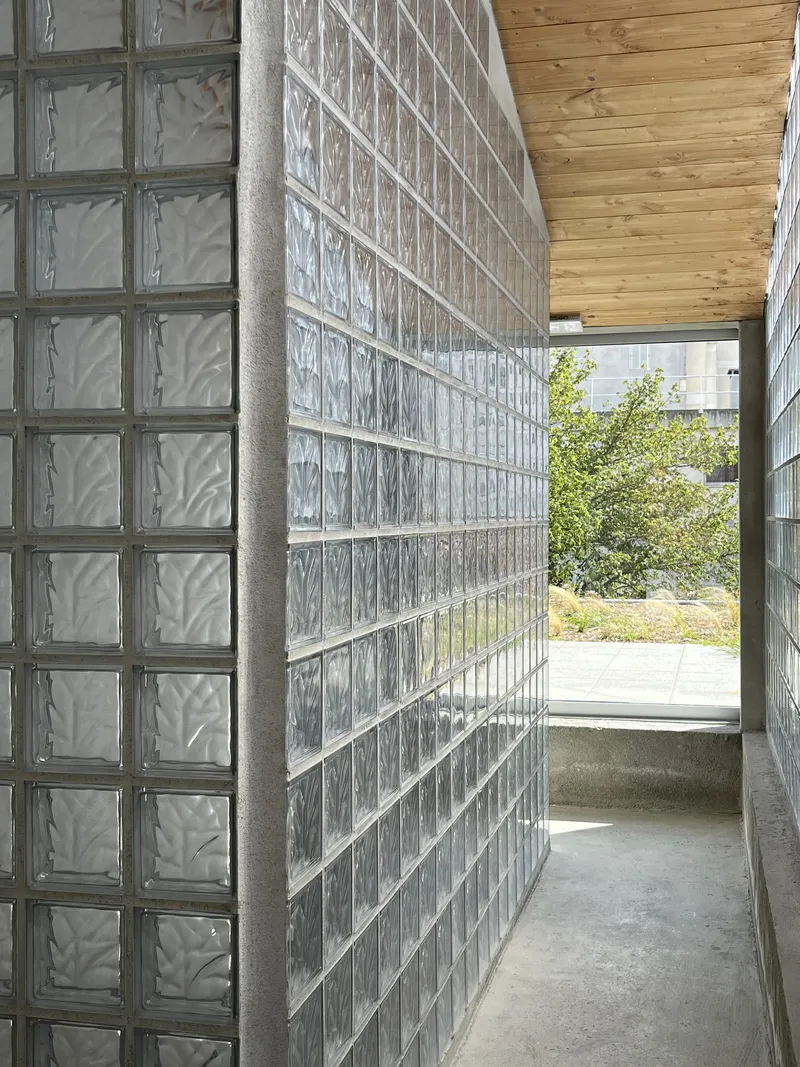 Corridor passage flanked by translucent glass block walls with timber ceiling opening to courtyard beyond