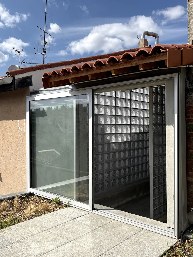 Rooftop terrace with sliding glass doors and curved glass block wall beneath terracotta tiles and blue sky