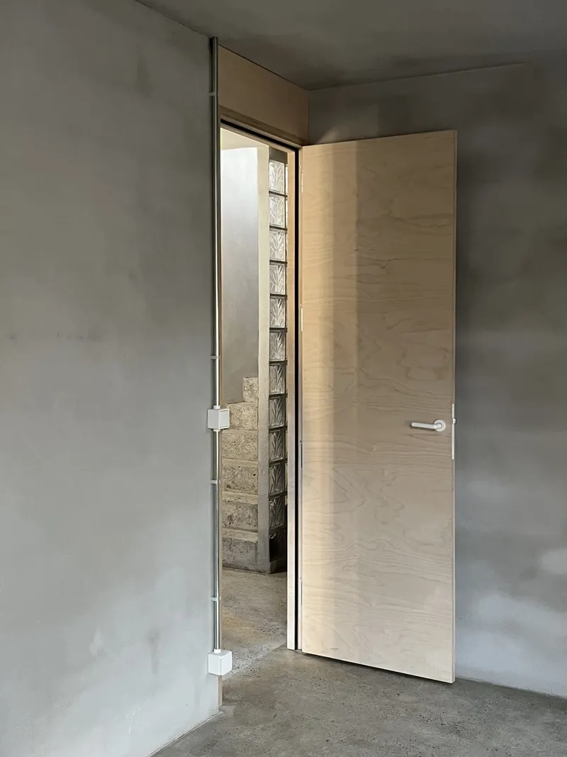 Light wood door partially open in concrete room revealing glass block wall in adjacent space