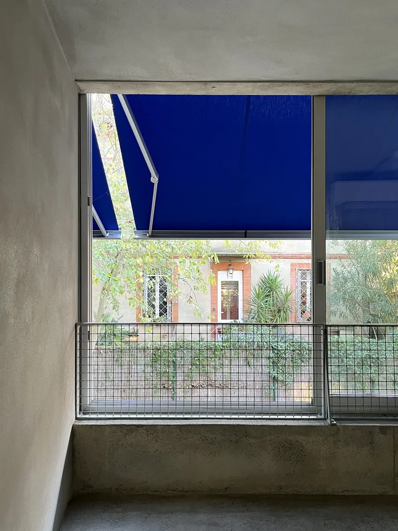 Window opening with blue retractable awning partially deployed above metal balustrade and planted garden