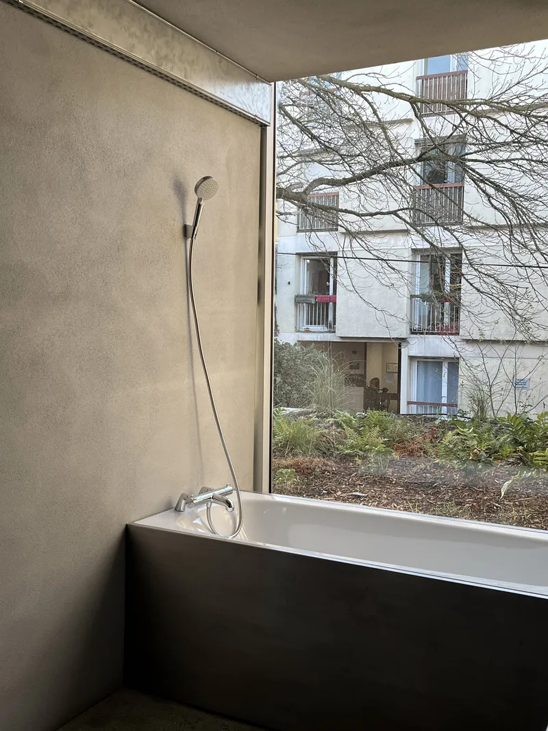 Bathtub positioned beneath corner window with view of bare trees and adjacent apartment facade