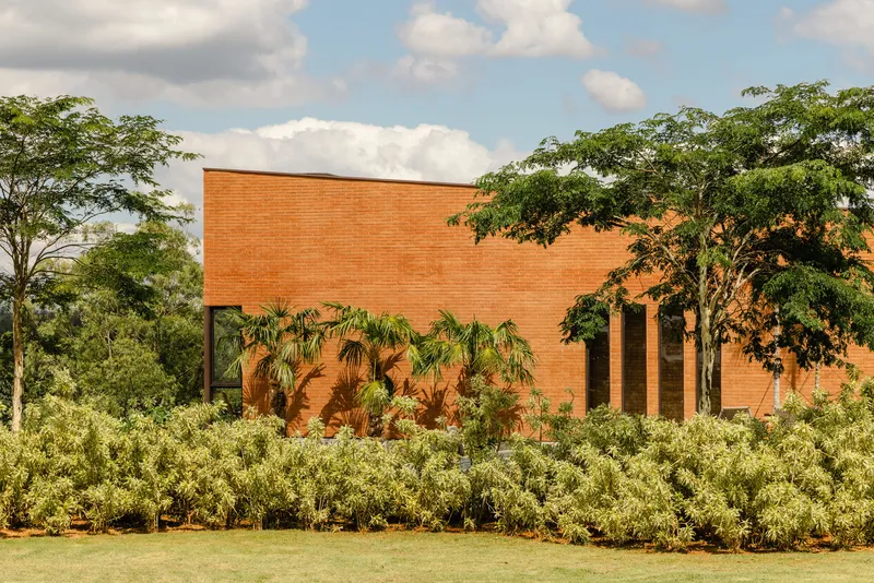 Red brick wall punctuated by tall glazed openings behind flowering shrubs and lawn