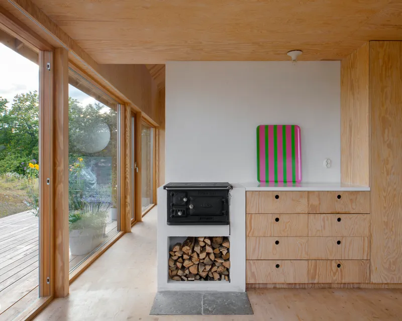 Cast iron wood stove with firewood storage niche beside plywood cabinetry under a timber ceiling