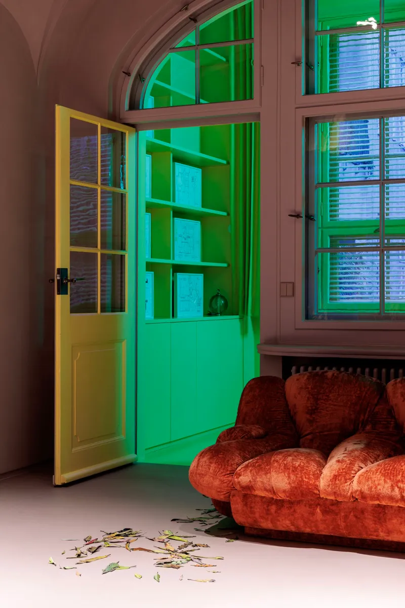 Green-lit alcove with open shelving viewed through yellow framed door beside arched window and velvet sofa