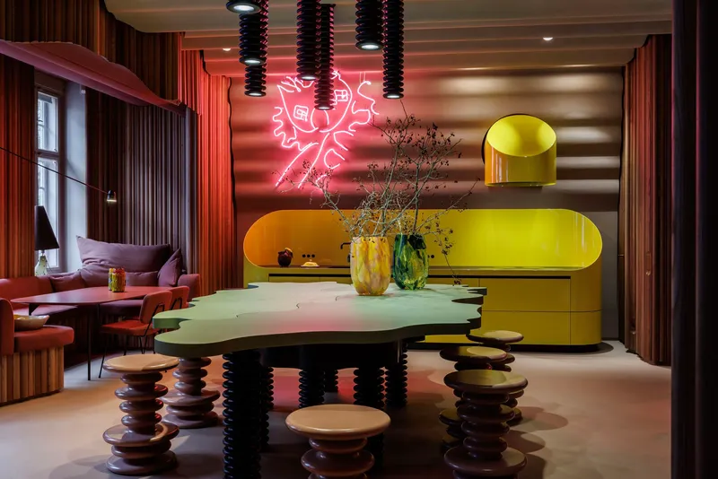Dining area with yellow curved cabinetry, neon wall art, and table on stacked disc base stools