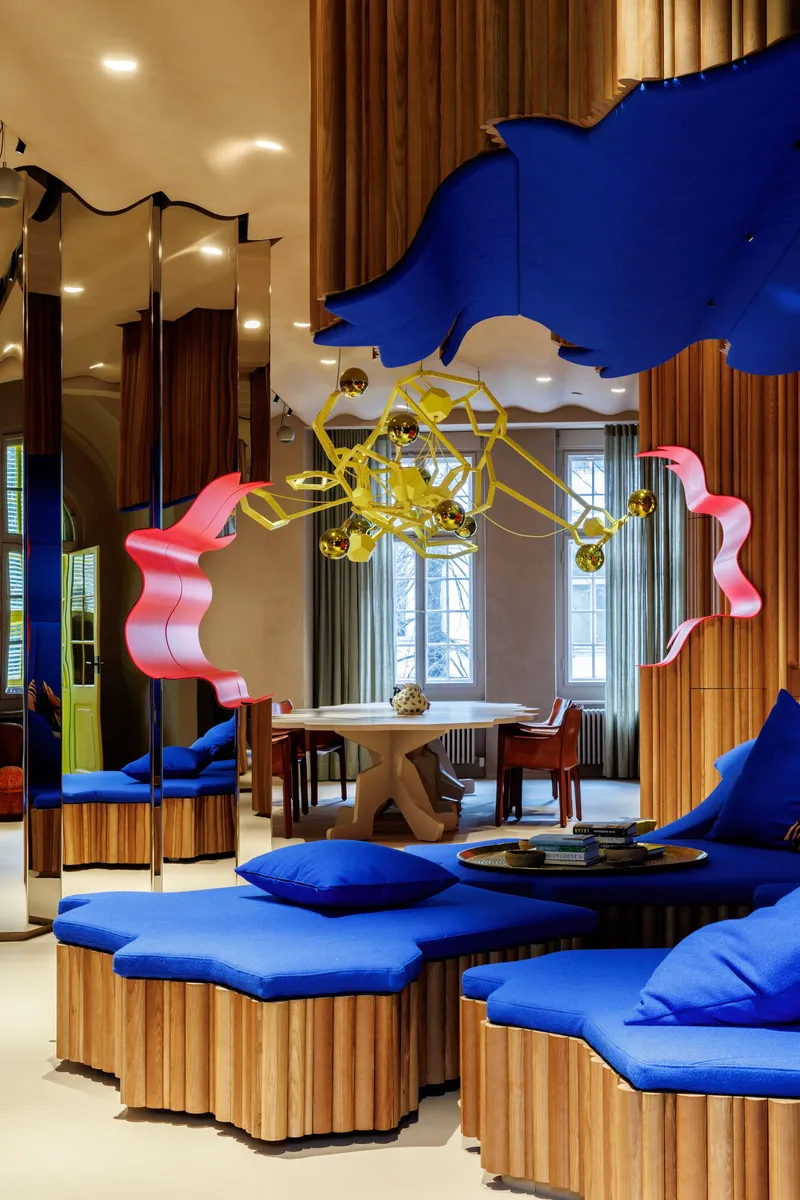 Seating area with blue upholstered benches on timber bases beneath a gold chandelier and hanging sculptures