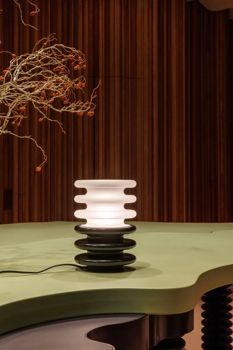 Stacked disc table lamp on a pale green surface against vertical timber paneling with dried branch overhead