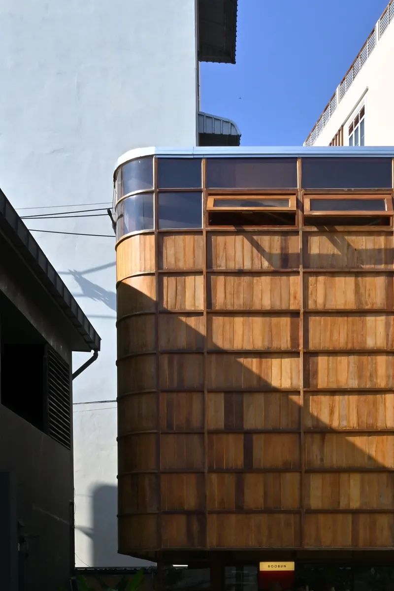 Curved timber-paneled facade with steel framing rising between adjacent buildings under clear blue sky