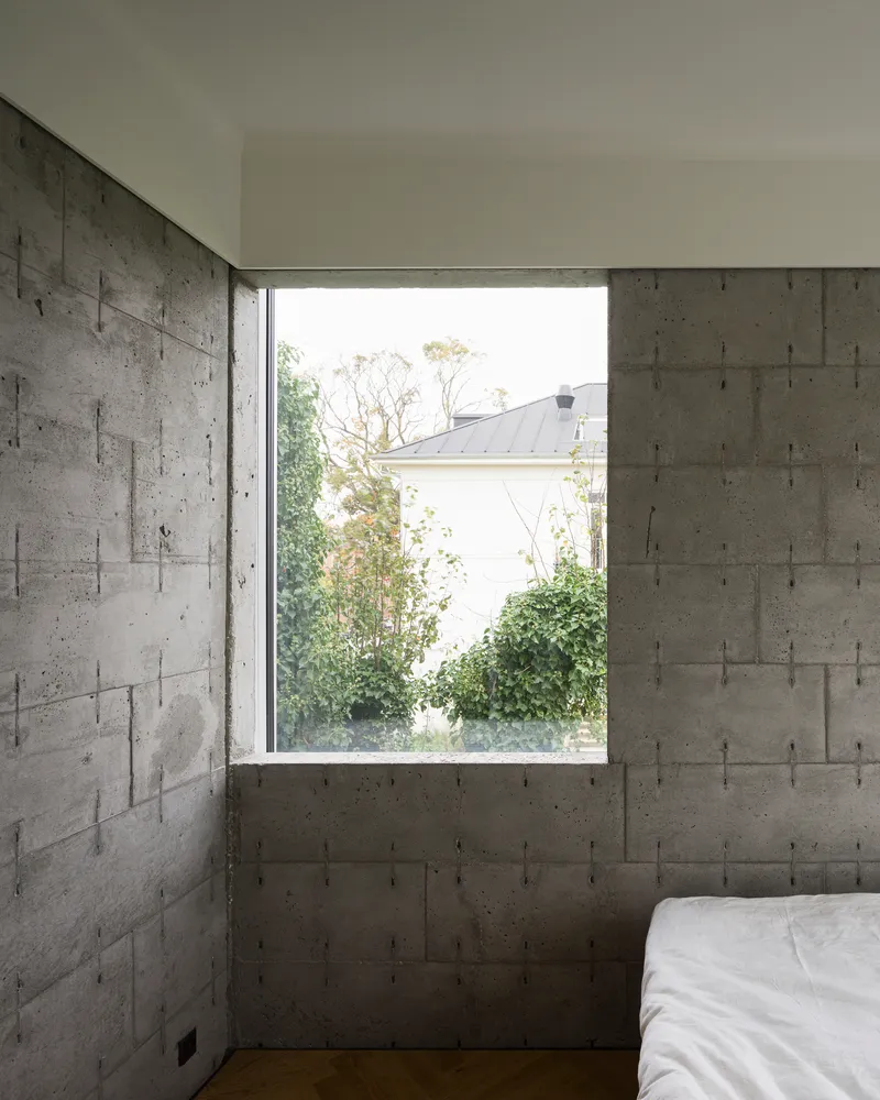 Bedroom corner with board-formed concrete walls and window overlooking trees and neighboring rooftop
