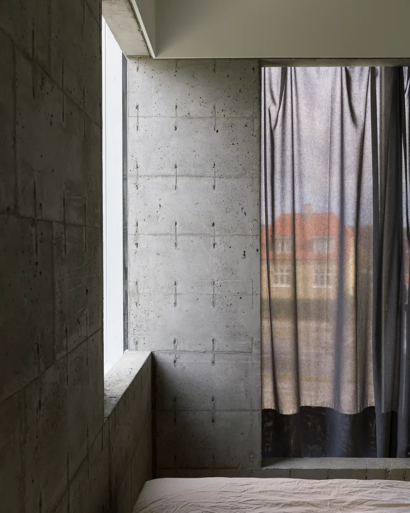 Bedroom corner with board-formed concrete walls, tall window slot, and draped curtain filtering soft daylight