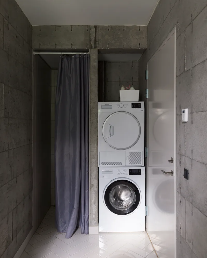 Compact laundry alcove with stacked washer and dryer flanked by dark curtains and concrete walls