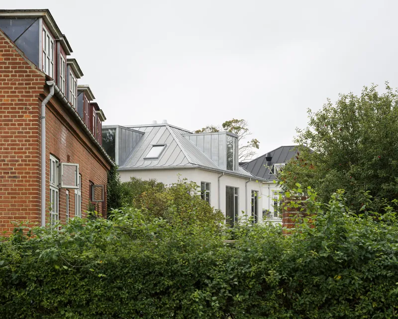 White house with standing seam metal roof nestled between brick buildings and dense hedgerow