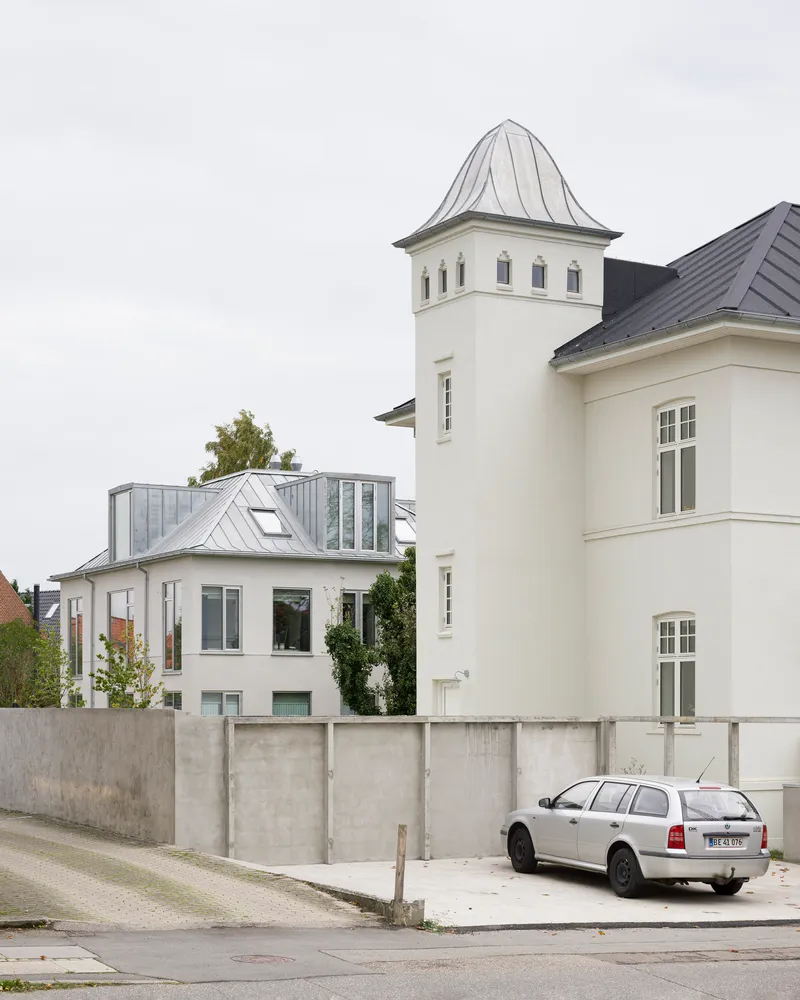 Street view of white tower with pointed roof beside zinc-clad addition behind concrete retaining wall