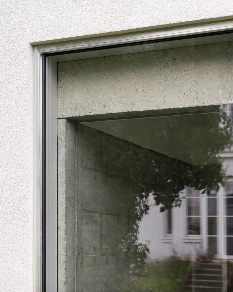 Window detail showing concrete lintel meeting plaster facade with reflected foliage in glass