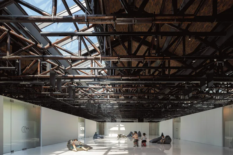 Gallery hall with exposed timber and steel truss structure above white exhibition walls and scattered visitors
