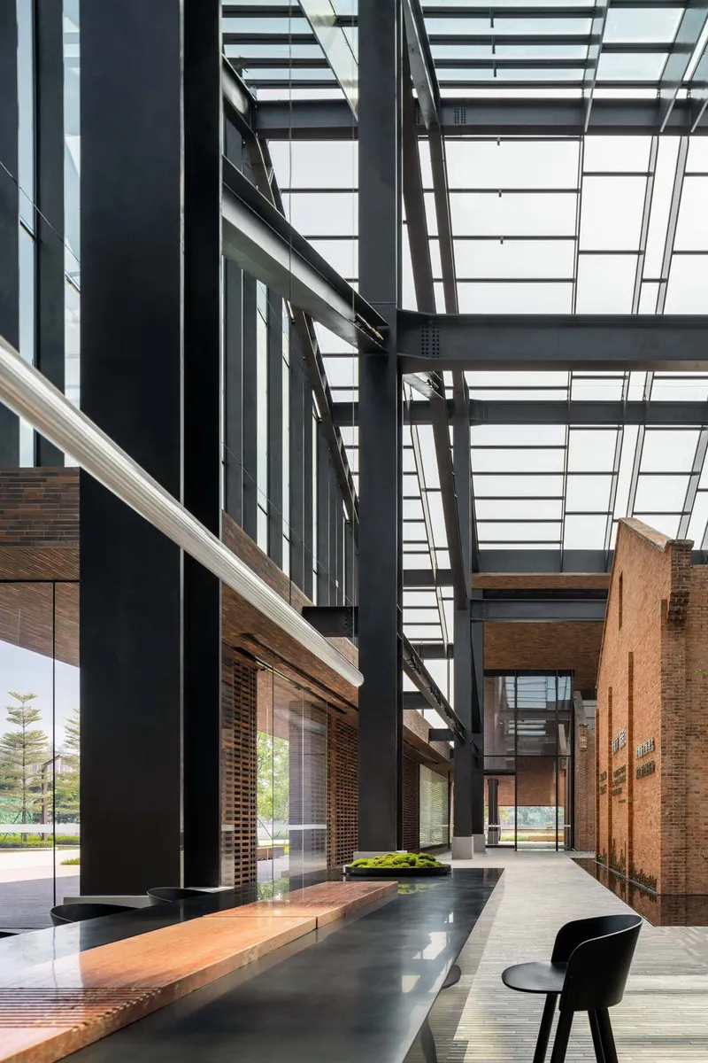 Atrium corridor with black steel structure and glazed roof above timber benches and planted beds
