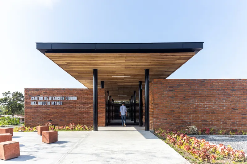Entry corridor with timber slat ceiling and black steel columns framing a figure walking toward the courtyard