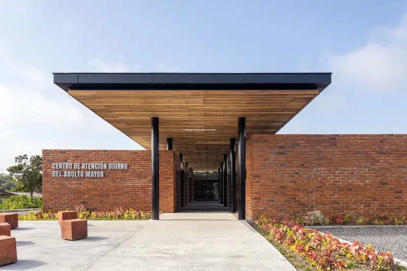 Entry facade with cantilevered timber soffit projecting over red brick wall and planted beds in daylight