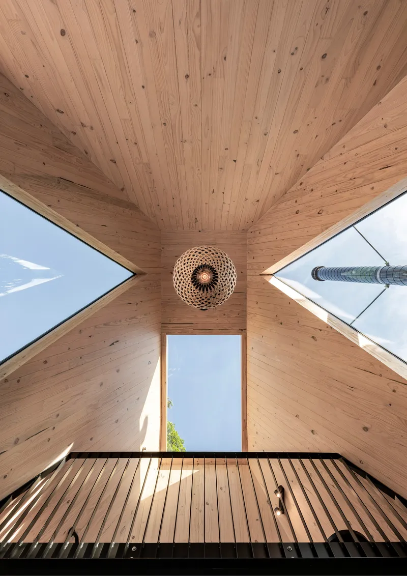 Upward view of timber-lined peaked ceiling with angled skylights and woven pendant centered above steel mezzanine railing