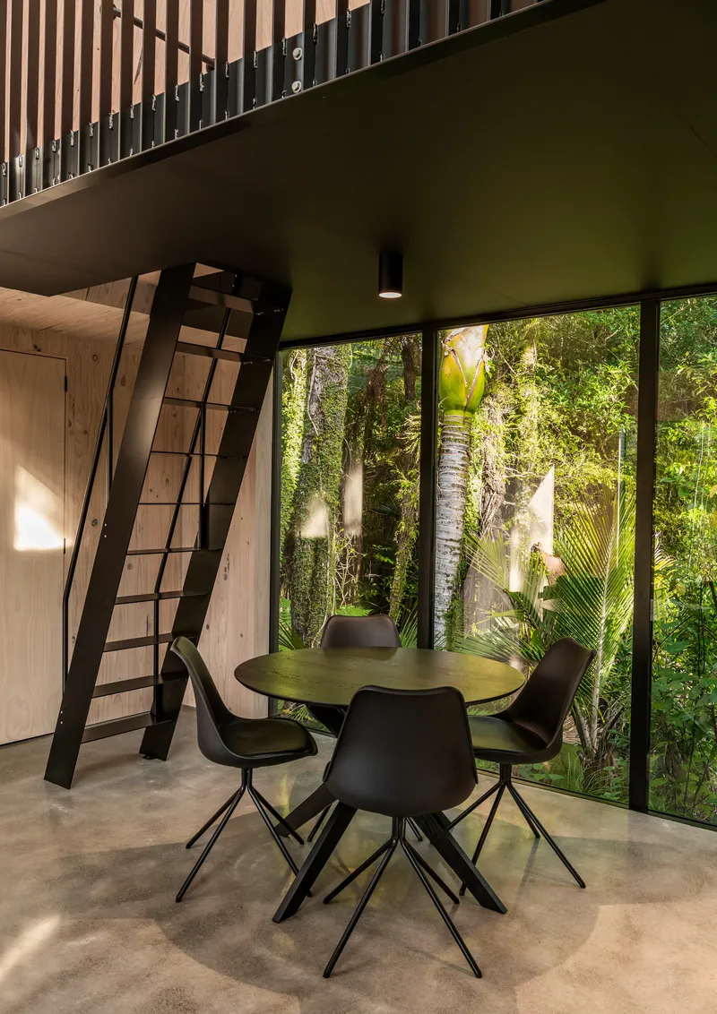 Dining area beneath mezzanine with ship ladder and full-height glazing framing palms in dappled afternoon light