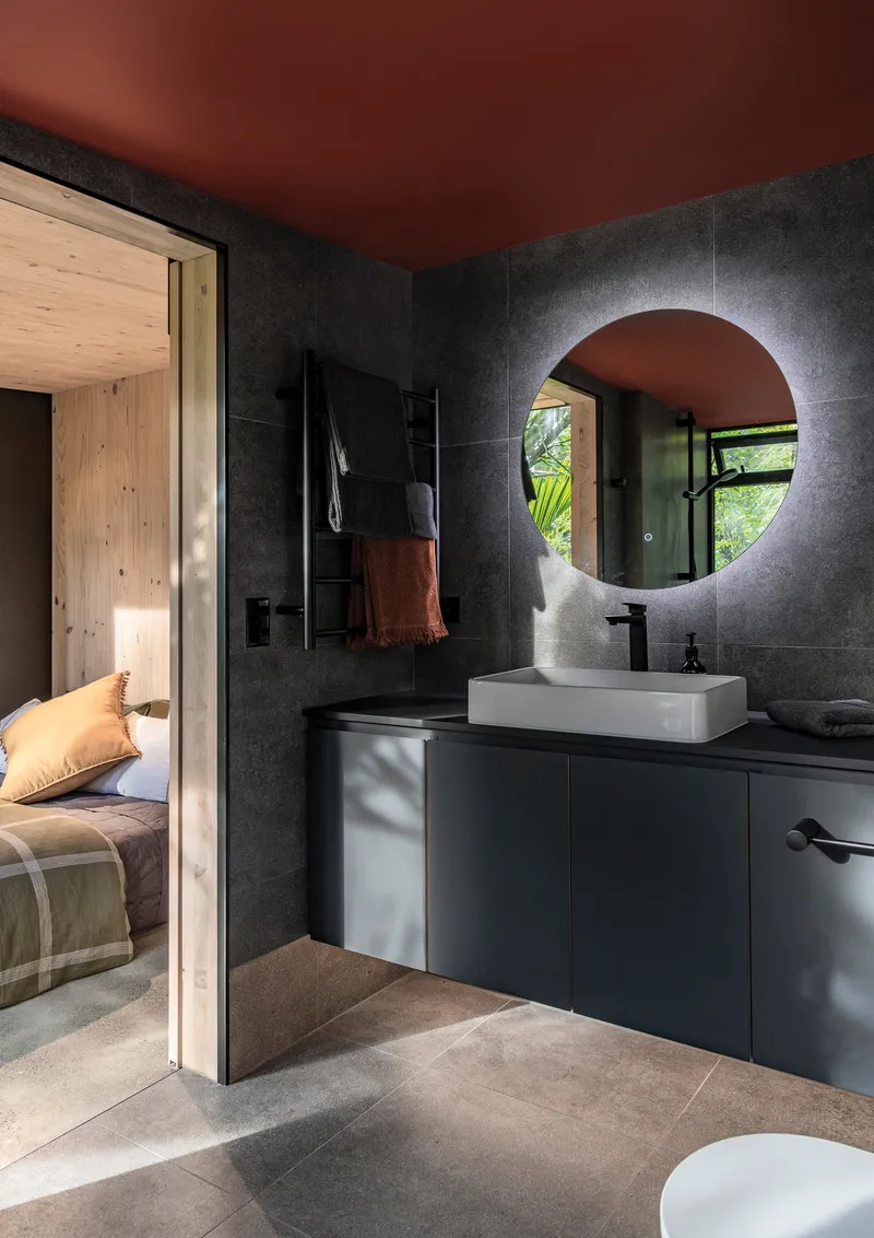 Bathroom with backlit round mirror and black tile walls opening to a timber-lined bedroom