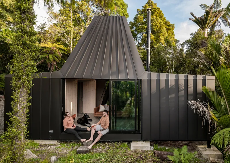 Black corrugated metal pavilion with conical roof and two people seated in open threshold beneath palms