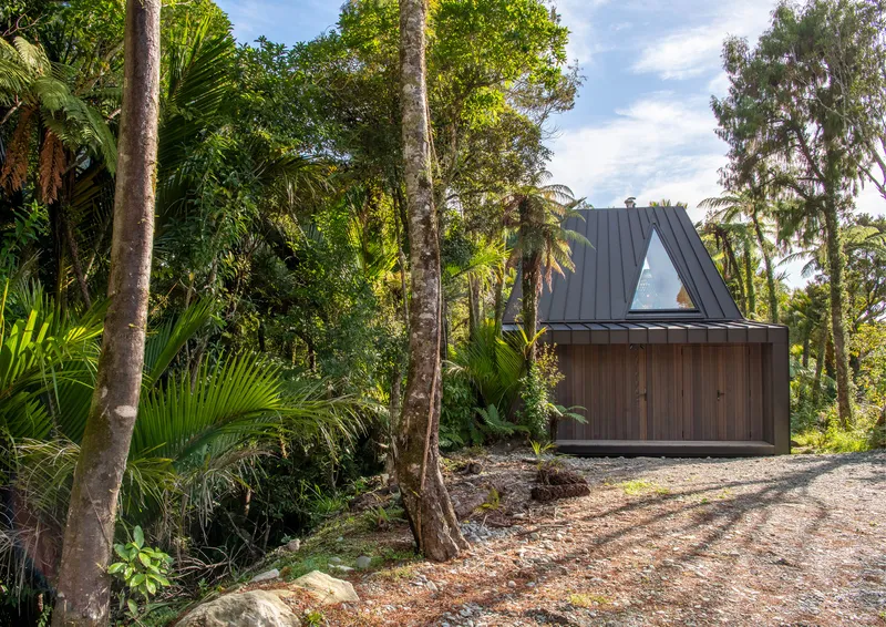 Timber pavilion with triangular window in metal roof accessed by gravel driveway through subtropical forest