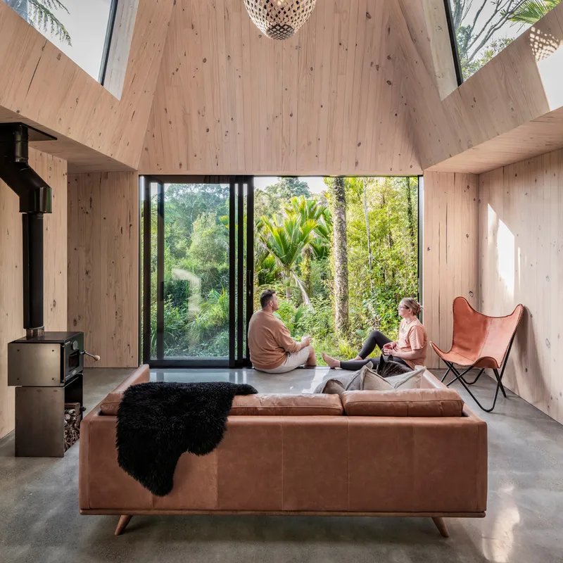 Interior with sloped plywood ceiling and two people seated on sofa facing full-height glazing to garden