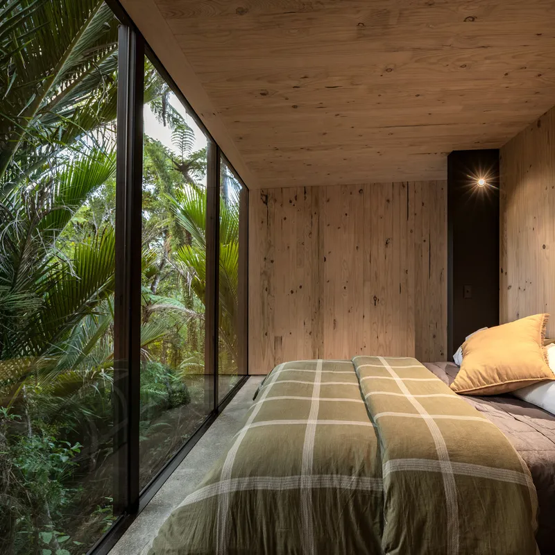 Sleeping area with plywood walls and ceiling and angled glazing overlooking palm fronds