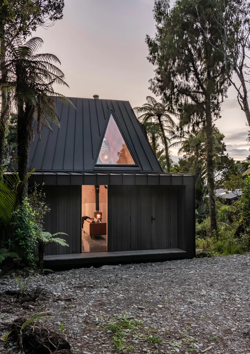 Steep-pitched black metal roof with triangular window glowing at twilight among tropical trees