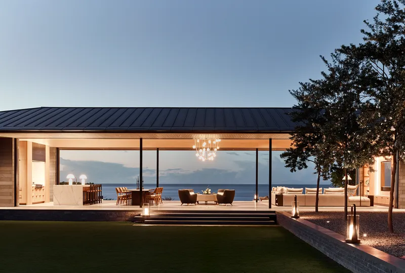 Glass-walled pavilion with standing seam metal roof opening to terrace and ocean view at twilight