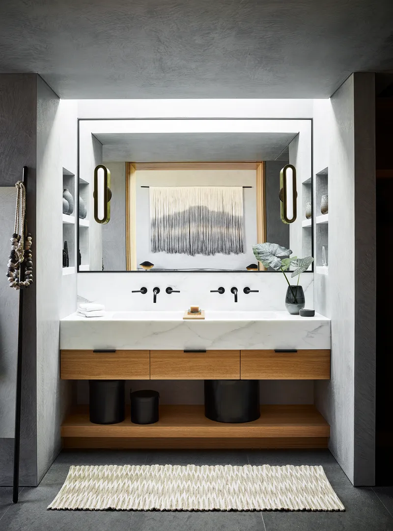 Bathroom vanity with floating stone counter, timber drawers, and oval sconces flanking recessed mirror