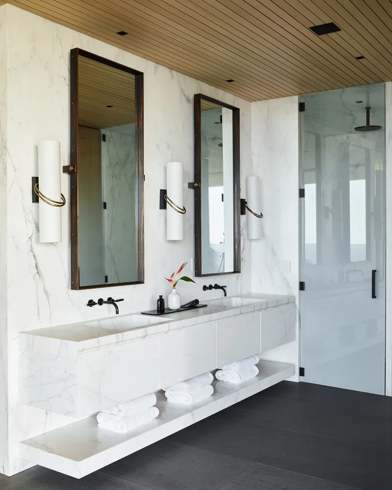 Marble-clad bathroom with twin mirrors, glass-enclosed shower, and slatted timber ceiling
