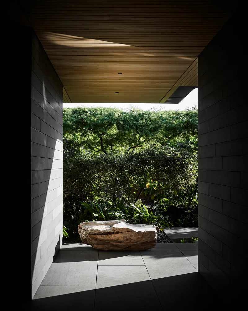 Covered passage with ribbed wood ceiling framing a sculptural boulder and dense tree canopy beyond