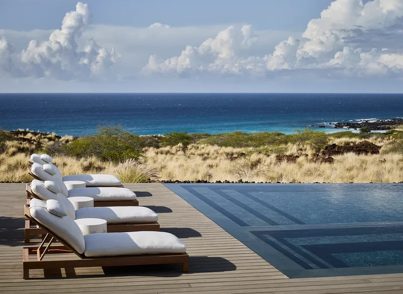 Timber deck with lounge chairs beside an infinity pool overlooking coastal grassland and ocean