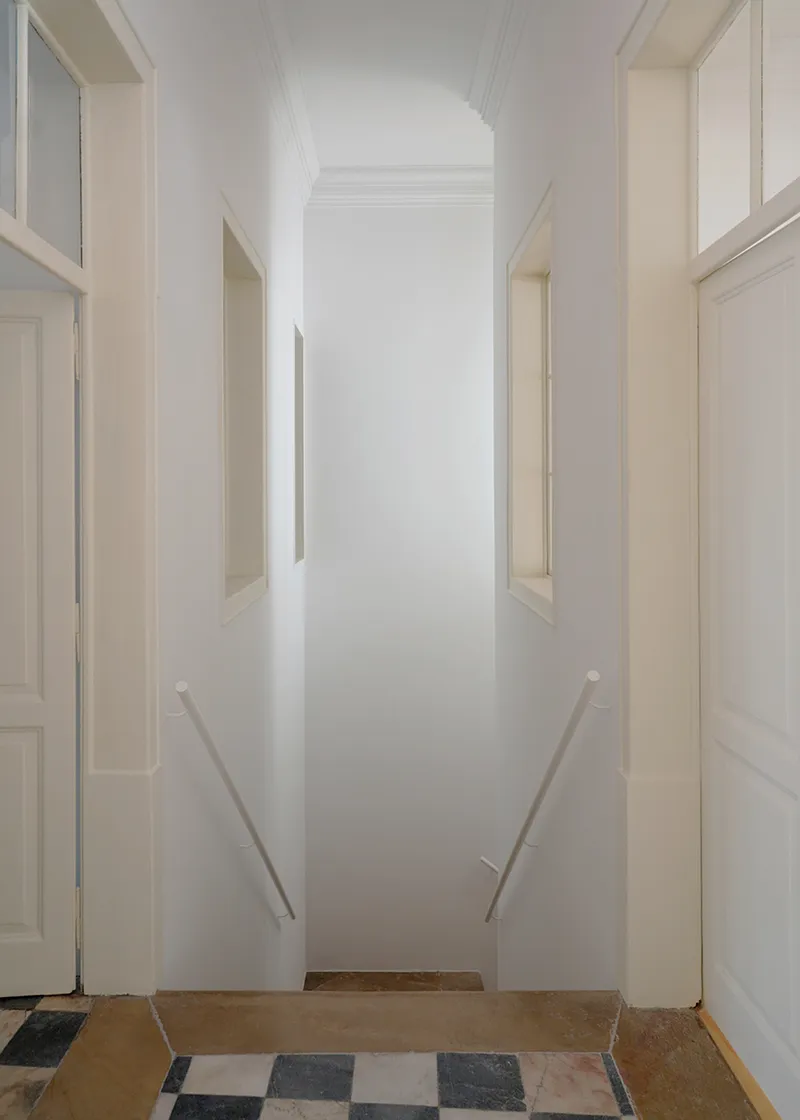 White corridor with vertical niches flanking walls and handrails leading to wooden step