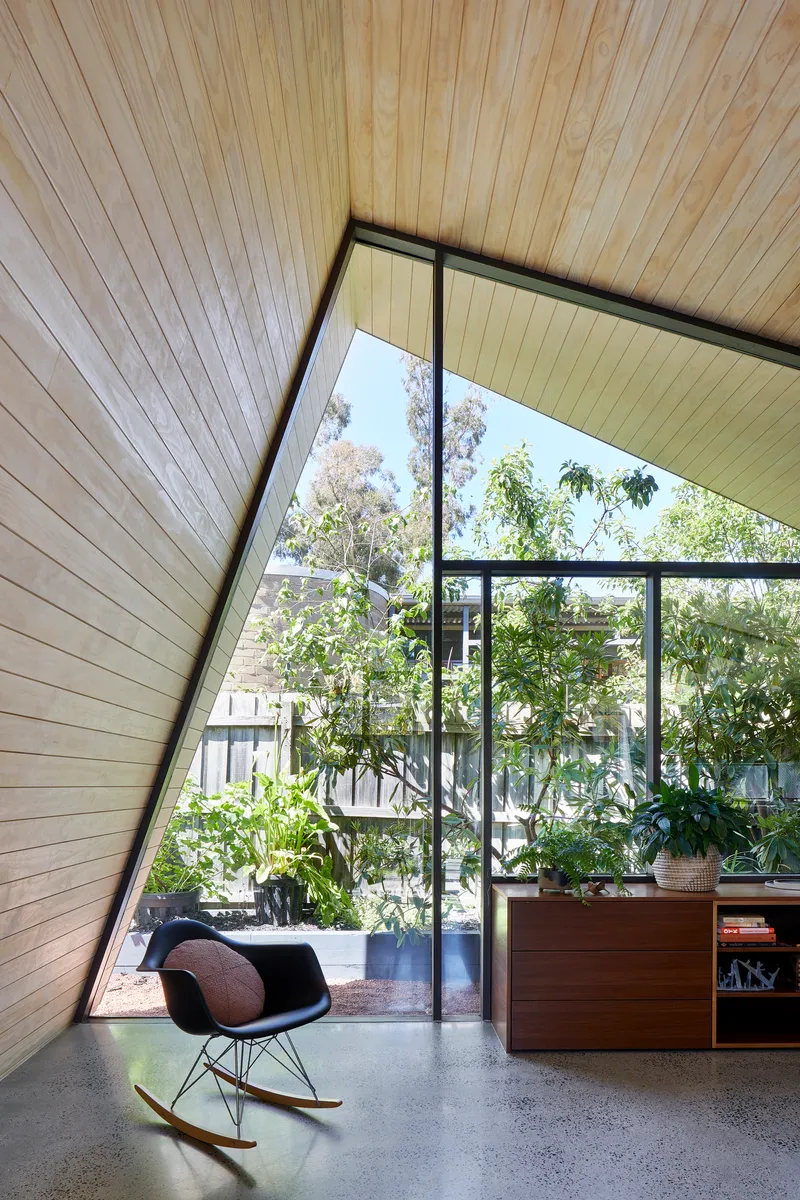 Corner room with raking timber ceiling and glazed wall overlooking a planted courtyard with rocking chair