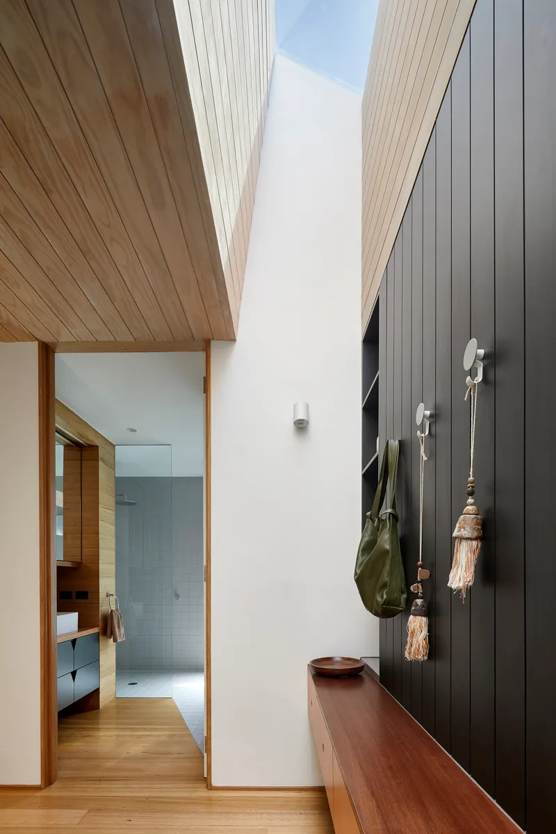Hallway with sloped timber ceiling panels converging above white walls and timber bench with hanging textiles