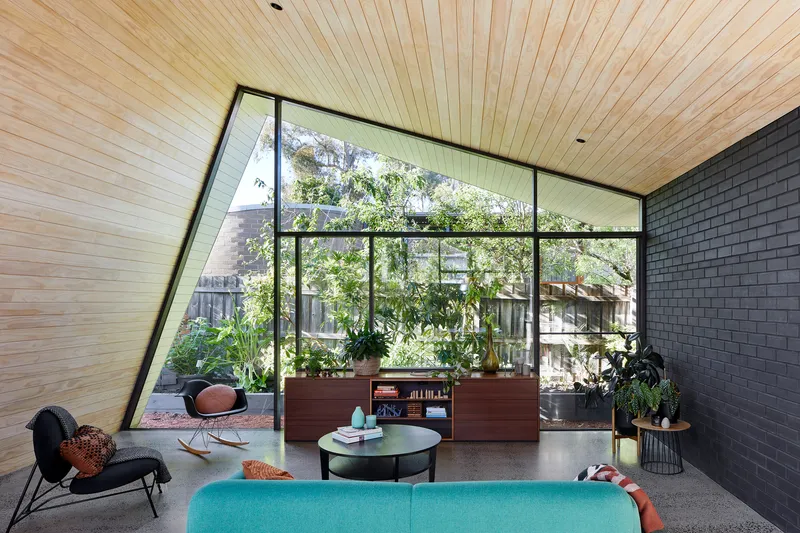 Living room with angled glazed wall framing garden views, timber plank ceiling and polished concrete floor