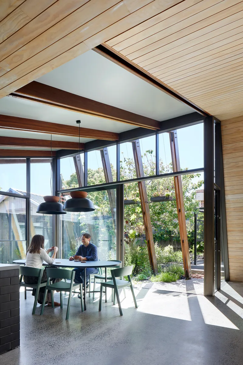 Dining space framed by angled timber columns visible through floor-to-ceiling glazing with two people conversing
