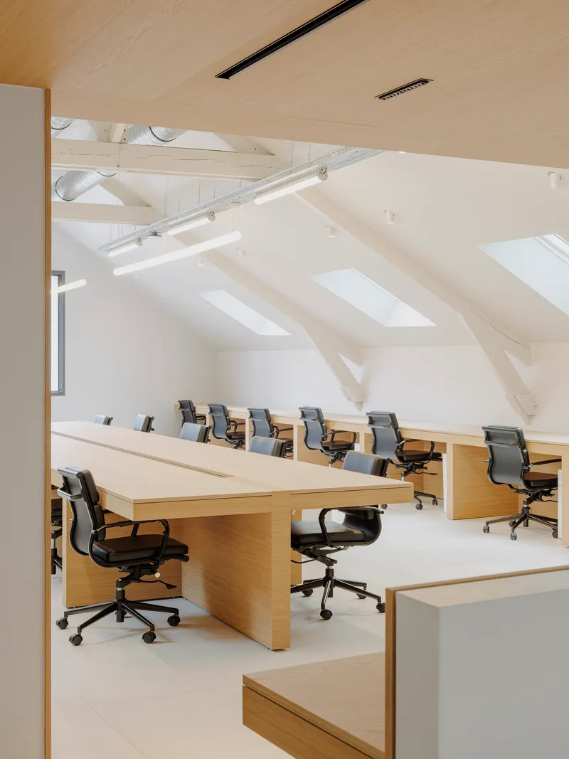 Open-plan office with rows of oak desks and office chairs under exposed white-painted roof trusses
