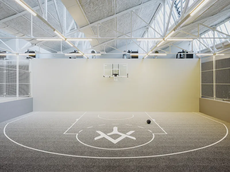 Indoor basketball court with white structural trusses overhead and tall glazed walls on both sides