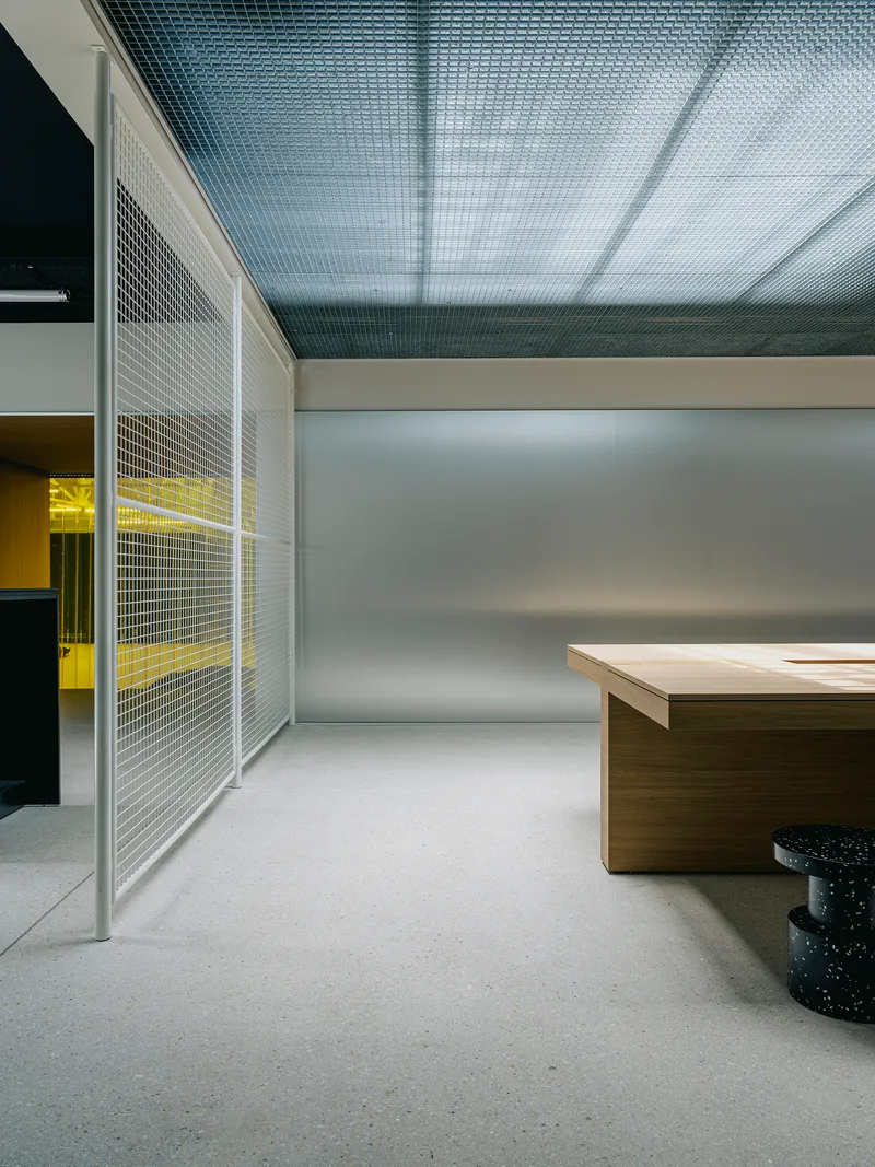 Interior space with metal mesh partition panels and yellow-lit room visible beyond timber desk